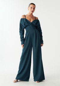 Tīrkīza krāsas kombinezons ar garām piedurknēm, plecus atverošu dizainu, drapētu augšdaļu, platu kāju biksēm un gludu auduma tekstūru. Sudraba siksniņu augstpapēžu kurpes.