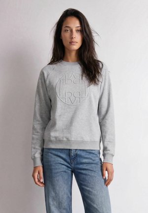 Kvinde med langt mørkt hår iført en lysegrå sweatshirt med præget rundt tekstdesign og blå jeans, stående mod en ensfarvet baggrund.