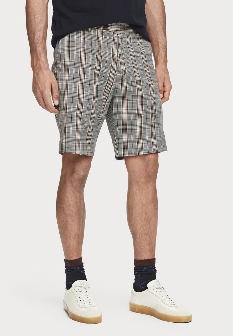 Homme portant un short gris à carreaux longueur genou, des baskets blanches avec semelles en gomme, des chaussettes foncées à motifs et une chemise sombre, debout sur un fond uni.