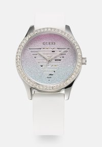 Rund Guess klokke med sølvfarge, hvit silikonrem, gradient dial med glitter fra rosa til blå, med trekantede utsparinger og krystall-accenter.