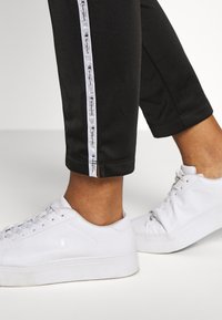 Svarta träningsbyxor med en vit logotyprand längs sidan, i kombination med vita sneakers. Byxorna har en kort längd och en figurnära design.