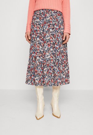 Femme portant une jupe midi fleurie bleu marine, un pull côtelé corail et des bottes blanches à bout pointu mi-mollet, debout devant un fond uni.