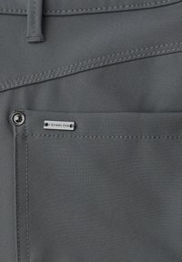 Grau Stoffhose mit detaillierten Nähten und einem kleinen silbernen Logoanhänger an der vorderen Tasche, mit glatter Textur und taillierter Passform.