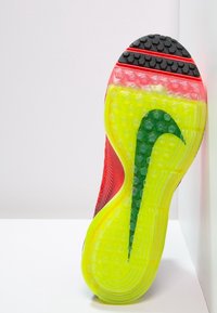 Nike teniska s texturovaným červeným svrškem, jasně žlutou podrážkou a zeleným swooshem. Disponuje hexagonálním vzorem na podrážce pro lepší přilnavost.