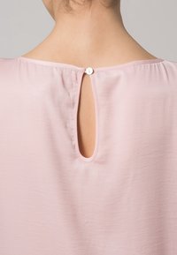 Arrière du cou d'une personne portant un chemisier rose clair avec une petite ouverture en trou de serrure fermée par un seul bouton blanc en haut.