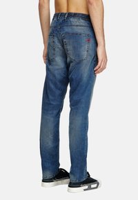 Pantalon en denim bleu avec une taille élastique, cinq poches et une finition délavée. Présente des coutures visibles et une étiquette logo rouge sur la poche arrière.