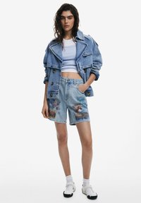 Jachetă din denim cu butoni dubli, top scurt ușor albastru și pantaloni scurți din denim cu modele multicolore și margini desfundate, purtate cu șosete și pantofi slip-on.