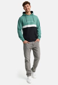 Felpa color block verde e blu navy con striscia bianca; tessuto misto cotone, cappuccio con cordino e polsini a coste. Indossata con jeans grigi e sneakers bianche.