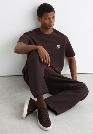 Homme assis sur un sol gris, portant un t-shirt marron foncé, un pantalon assorti, des chaussettes et des baskets marron avec des semelles blanches, contre un mur blanc.