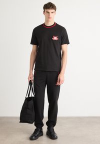 Zwart katoenen T-shirt met rode ribbelhals en geborduurde graphic. Gecombineerd met zwarte joggers en zwarte sneakers. Houdt een zwarte tote bag vast.