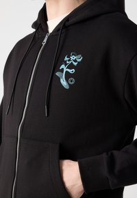 Sweat à capuche zippé noir avec un dessin de fleur stylisée bleu clair et un visage souriant sur la poitrine, porté avec une main dans la poche.