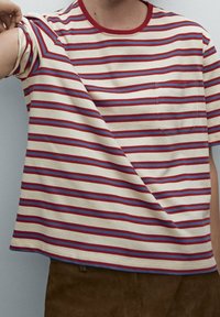 Henkilö pukeutuneena kermaan vivahtavaan t-paitaan, jossa punaisia ja sinisiä vaakaraitoja, rintatasku, sekä ruskeat housut, nostaa toista kättään hieman sivulle.
