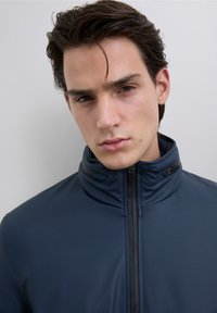 Mörkblå jacka med hög krage och dragkedjestängning. Slät, lättviktsmaterial med kontrasterande svarta detaljer på kragen.