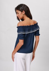 Top blu navy a spalle scoperte con scollatura a volant, caratterizzato da bordi decorativi bianchi e maniche a palloncino corte. Abbinato a pantaloni bianchi.