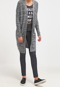 Długi, otwarty kardigan z przodu wykonany z melanżowego czarno-białego dzianiny, w połączeniu z czarnym T-shirtem z nadrukiem i ciemnymi dżinsami. Kardigan posiada ściągacze na mankietach i dole.