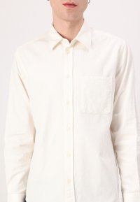 White corduroy shirt with a pointed collar, long sleeves, front button closure, and a chest pocket on the left side. Subtle ribbed texture.