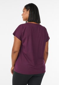 Frau mit schulterlangen, glatten Haaren, die sich abwendet, trägt ein kurzärmeliges lila Shirt und schwarze Hosen vor einem schlichten Hintergrund.