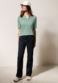 Polo shirt verde chiaro a maniche corte, jeans dritti blu scuro, cappellino bianco e sneakers crema con accenti chiari. Outfit semplice e casual.