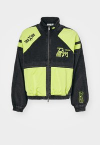 Svart och neongrön Adidas zip-up jacka med texten "TEAM" och "TRAXXION", elastiska manschetter och midjeband samt hög krage mot vit bakgrund.