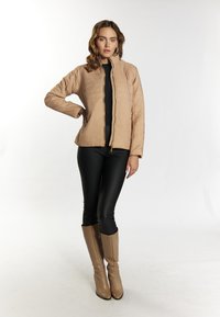 Beige geïsoleerde jas met een ritssluiting aan de voorkant, hoge kraag en geribbelde textuur, gecombineerd met zwarte leggings en beige laarzen tot aan de knie.