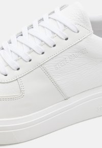 Zapatilla de cuero blanca con panel lateral texturizado, cordones blancos planos y logo en relieve. Incluye una suela de goma y detalles de costura mínimos.