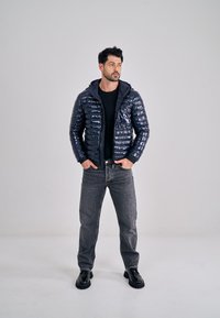 Marineblauwe pufferjack met glanzende afwerking, ritssluiting en capuchon, gedragen over een zwart shirt en grijze jeans. Zwarte laarzen met een dikke zool.