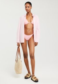 Różowy bikini składający się z trójkątnego stanika i majtek o niskim stanie, noszony pod jasnoróżową koszulą w paski. Akcesoria obejmują plecioną torbę i sandały.