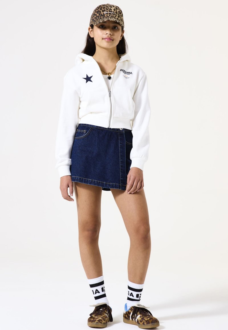 Sudadera blanca recortada con un gráfico de estrella, falda de mezclilla oscura, gorra de estampado de leopardo y zapatillas marrones con tiras. Calcetines con un patrón blanco y negro.