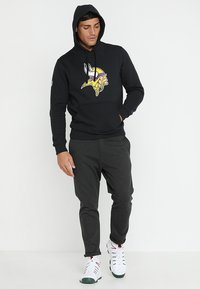 Sudadera negra con el logo de los Vikings, hecha de una mezcla de algodón, con un bolsillo canguro y puños acanalados. Combinada con pantalones gris ajustados y zapatillas blancas.