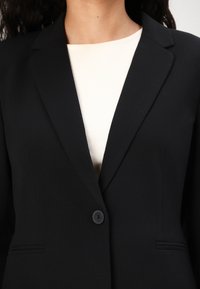 Svart skreddersydd blazer med klipp med notcher, enkle knapplukking, og sidelommer, båret over en kremfarget ribbestrikket topp. Glatt stoffstruktur.