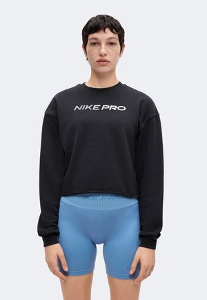 Kvinde iført en sort langærmet Nike Pro sweatshirt og lyseblå Nike Pro cykelshorts med høj talje står mod en ensfarvet baggrund.