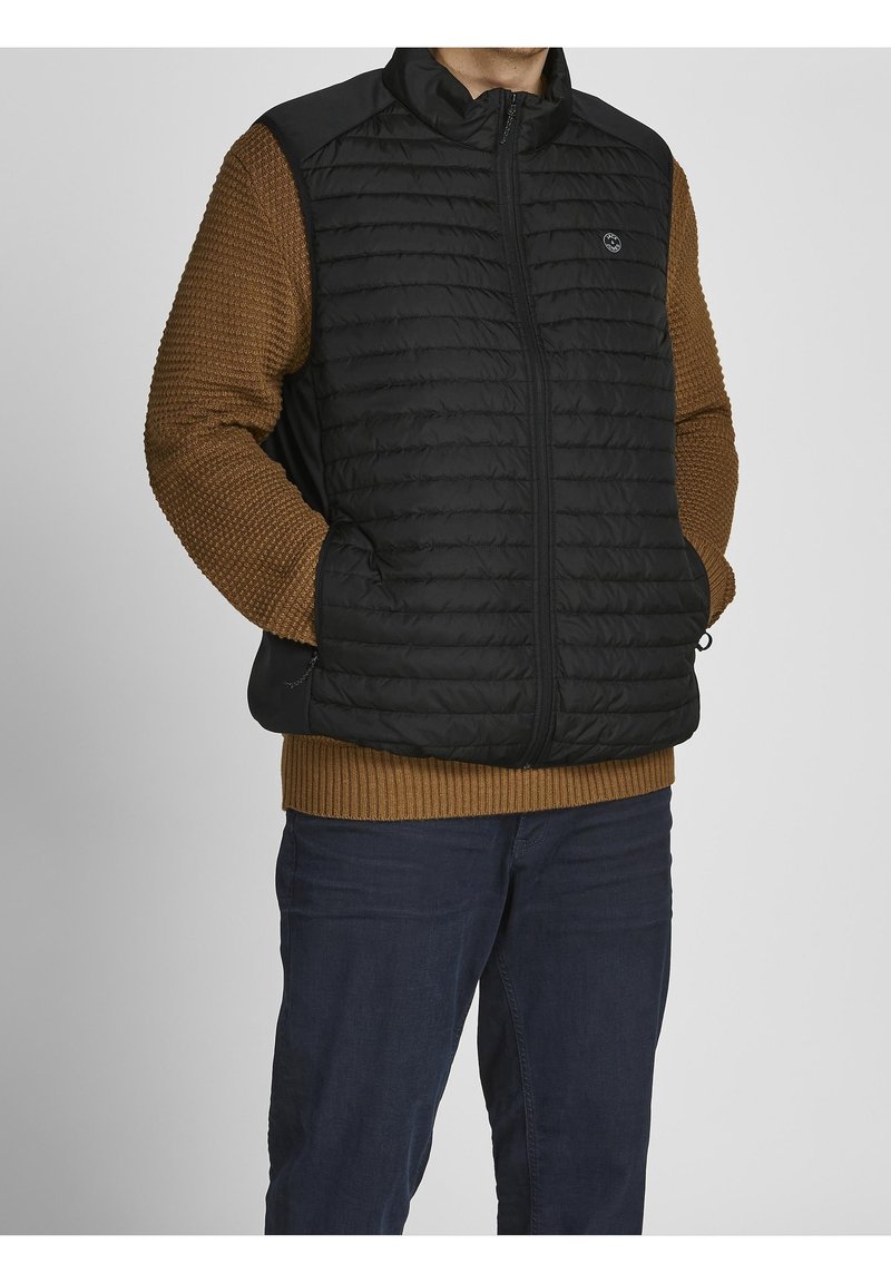 Czarna puchowa kamizelka z poziomym pikowaniem, zapinana na zamek błyskawiczny, z wysokim kołnierzem, zestawiona z brązowym swetrem o fakturze i niebieskimi dżinsami.