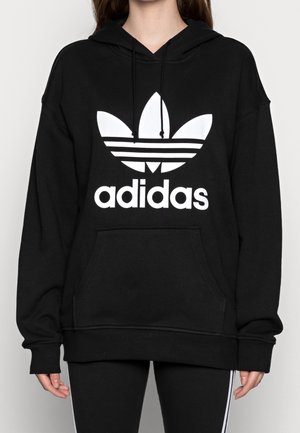 Personne portant un sweat à capuche noir Adidas avec un grand logo trefoil blanc et du texte sur le devant, assorti à un pantalon noir avec des bandes blanches sur les côtés.