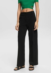 Pantalons noirs à jambe large avec une coupe soignée, en tissu lisse, et un unique bouton à l'avant. Associés avec un crop top côtelé vert.