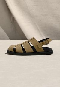 Sandale pour homme en daim marron clair, style pêcheur, avec semelle noire et bride arrière ajustable, présentée sur une surface texturée neutre.