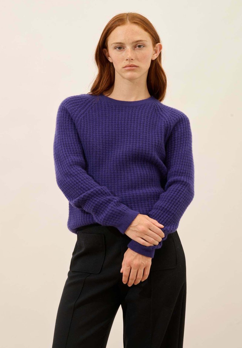 Violetti neulepusero, jossa on tekstuuri, ribbaatut hihansuut ja pyöreä pääntie. Yhdistetty mustiin housuihin, jotka korostavat rentoa istuvuutta.