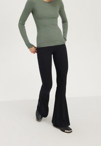 Olivgrünes, langärmeliges, geripptes Oberteil kombiniert mit schwarzen, ausgestellten Leggings, die eine glatte Textur und eine enge Passform aufweisen. Schwarze Schuhe mit silbernen Akzenten.