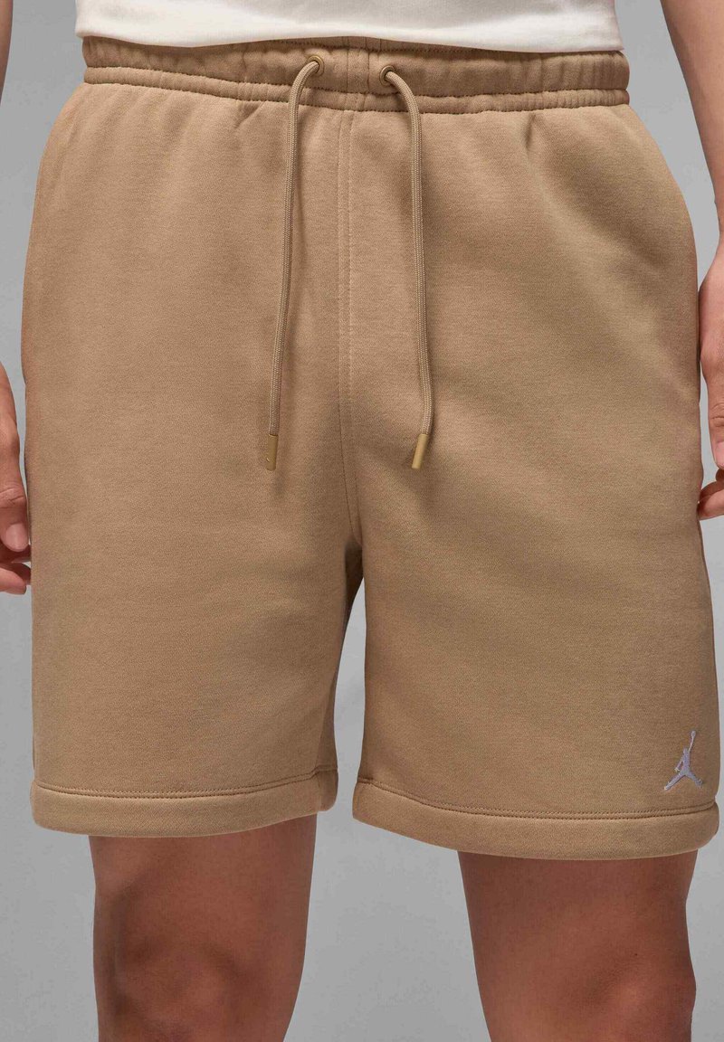 Shorts en coton beige avec une taille élastique, fermeture à cordon, et un petit logo blanc en bas à gauche. Texture lisse.