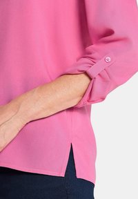 Rosa transparente Bluse mit hochgerollten Ärmeln und einem kleinen Knopfdetail. Seitliche Schlitze am Saum. Weiche Stoffstruktur.