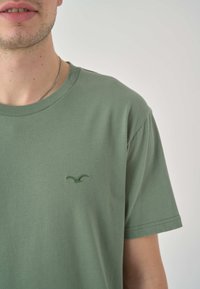 Grünes Baumwoll-T-Shirt mit kurzen Ärmeln, Rundhalsausschnitt und einem kleinen gestickten Logo auf der linken Brust, das eine glatte Textur und ein lässiges Design aufweist.