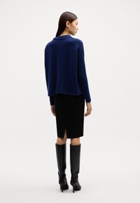 Dunkelblauem Wollmischpullover mit lockerer Passform, kombiniert mit einem schwarzen Bleistiftrock mit Schlitz und knielangen schwarzen Stöckelschuhen.