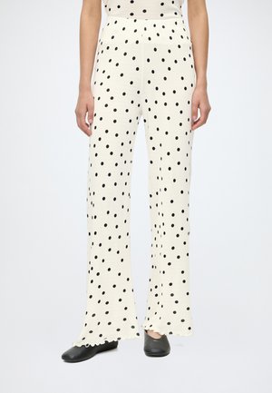 Femme portant un pantalon blanc taille haute avec des pois noirs, ourlets évasés, assorti à des chaussures noires sans lacets, debout sur un fond blanc.