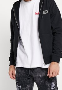 Felpa nera con zip realizzata in tessuto morbido, dotata di tasca frontale e discreto logo. Abbinata a una t-shirt bianca e pantaloni della tuta a fantasia.