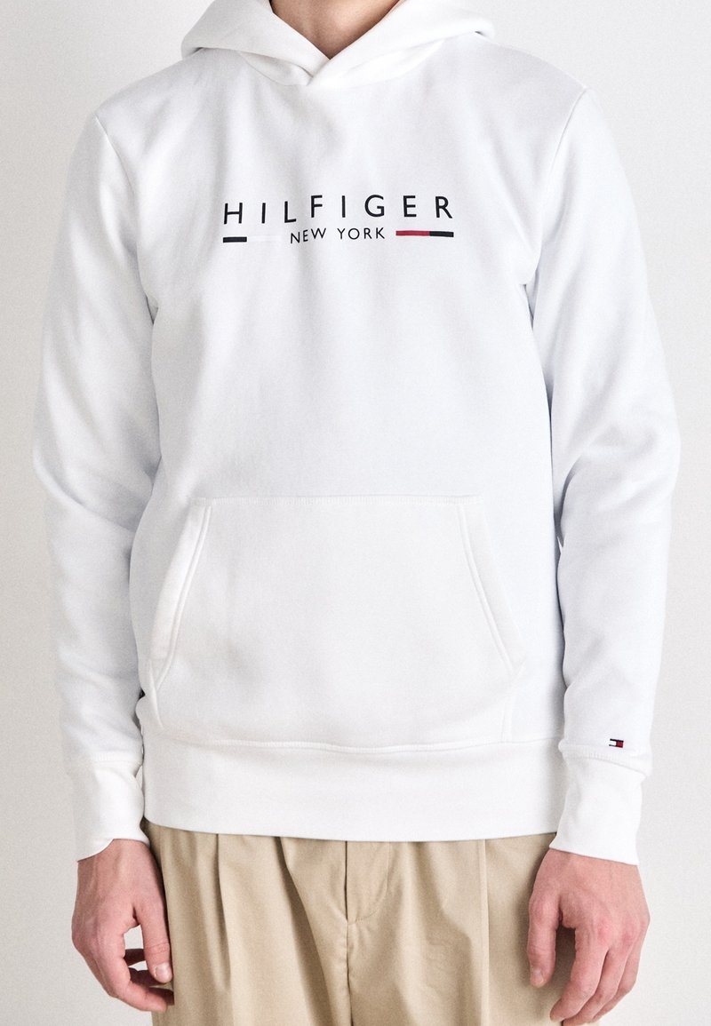 Sweat à capuche blanc avec le texte "HILFIGER NEW YORK" et des rayures rouges, blanches et noires, porté par une personne avec un pantalon beige.