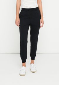 Calças de jogging pretas em material macio e elástico. Apresentam cós elástico, tornozelos com punhos e bolsos laterais. Usadas com ténis brancos.