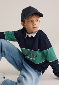 Laivastonsininen ja vihreä raidallinen pitkähihainen poolopaita, jossa on valkoinen kaulus ja teksti "NORTELD League", sekä vaaleansiniset farkut.