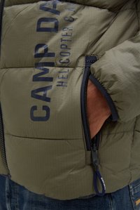 Mano nella tasca della giacca imbottita verde oliva con cerniera nera e testo blu scuro "CAMP DAVID HELICOPTER CA" sulla giacca.