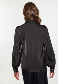Blusa nera in satin con maniche lunghe a sbuffo, polsini abbottonati e una texture liscia e lucida. Presenta un colletto classico e una vestibilità sartoriale.