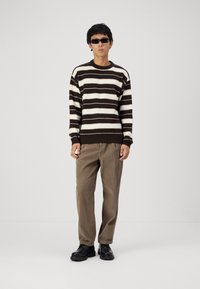 Brown and cream striped knit sweater with a crew neck, paired with loose tan corduroy trousers and black chunky shoes. Wearing black sunglasses.