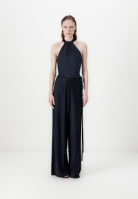 Navy pleated halter jumpsuit with a fitted bodice and wide legs. Features thin straps and a side tie for adjustable fit. Smooth texture.